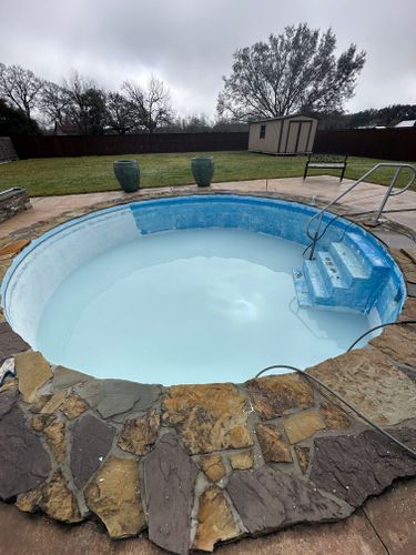 A swimming pool with stairs surrounded by rocks in a backyard.