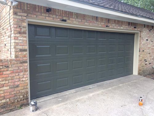 A garage door is being painted black in front of a brick building.