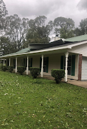 A white house with a green roof and a porch is sitting on top of a lush green field.