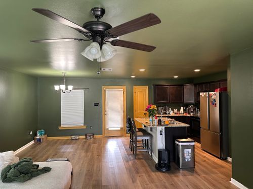A living room with a ceiling fan and a kitchen in the background.