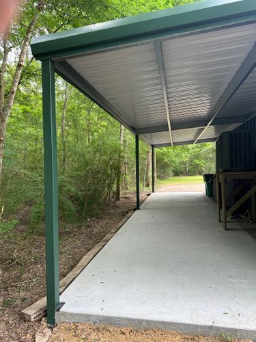 A carport with a green roof and a concrete walkway in the woods.