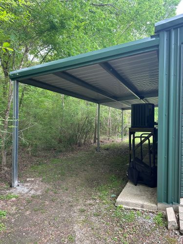 A green metal shed with a covered parking area in the woods.