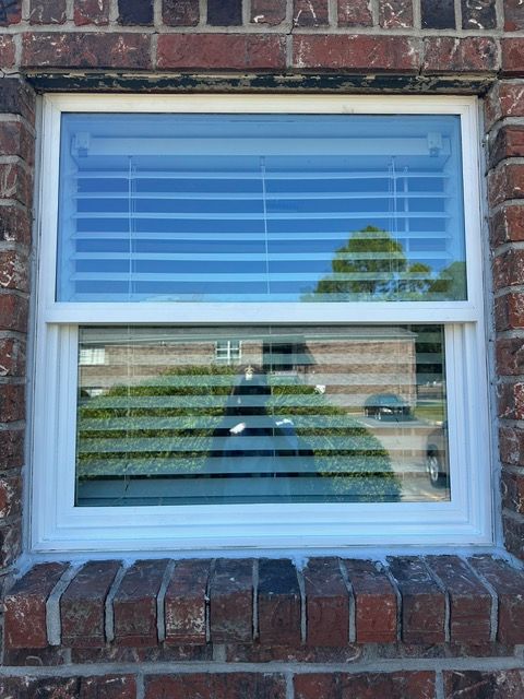 White-framed window in a red brick wall, blinds partially open, reflecting a building and trees.