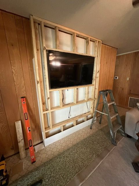 Wooden frame built around a mounted TV on a paneled wall, construction in progress with a ladder and level nearby.