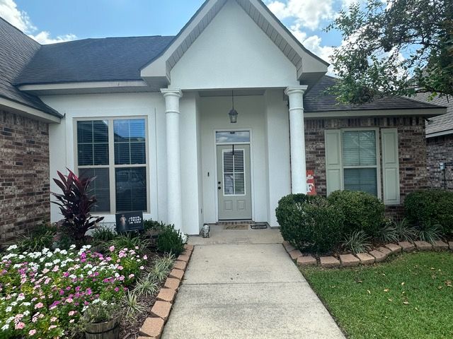 A white-painted house with columns, a front door, and a walkway surrounded by flowers and greenery.