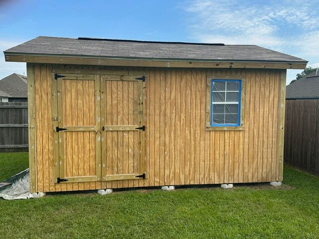 Wooden shed with double doors, blue-framed window, and gray roof, sitting on concrete blocks on a green lawn.
