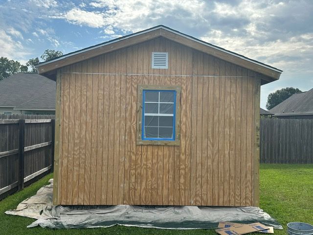 Wooden shed in backyard with blue-taped window and small vent.  Gray tarp on the ground, green grass.