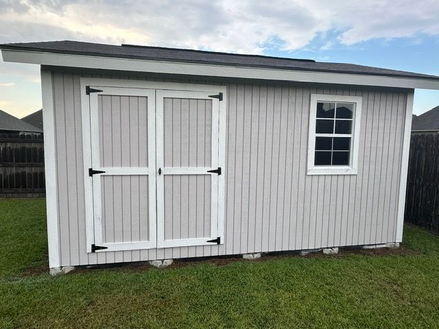 Gray shed with white-trimmed doors and window, black hinges, on a grassy lawn with a cloudy sky.