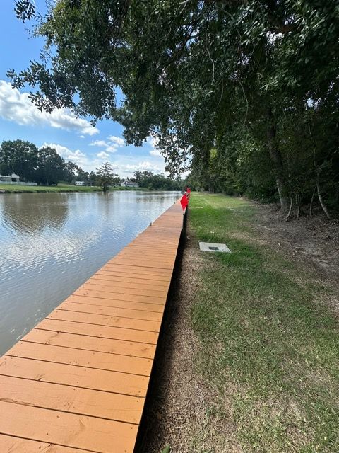 Wooden dock extends over water, grassy bank to the right. Person in red stands near the end. Sunny day.