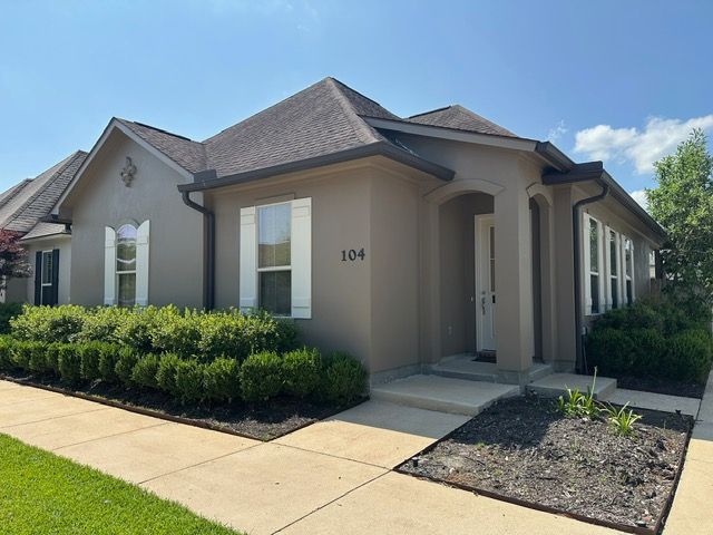Tan stucco house with white shutters, black trim, and green bushes. Address 
