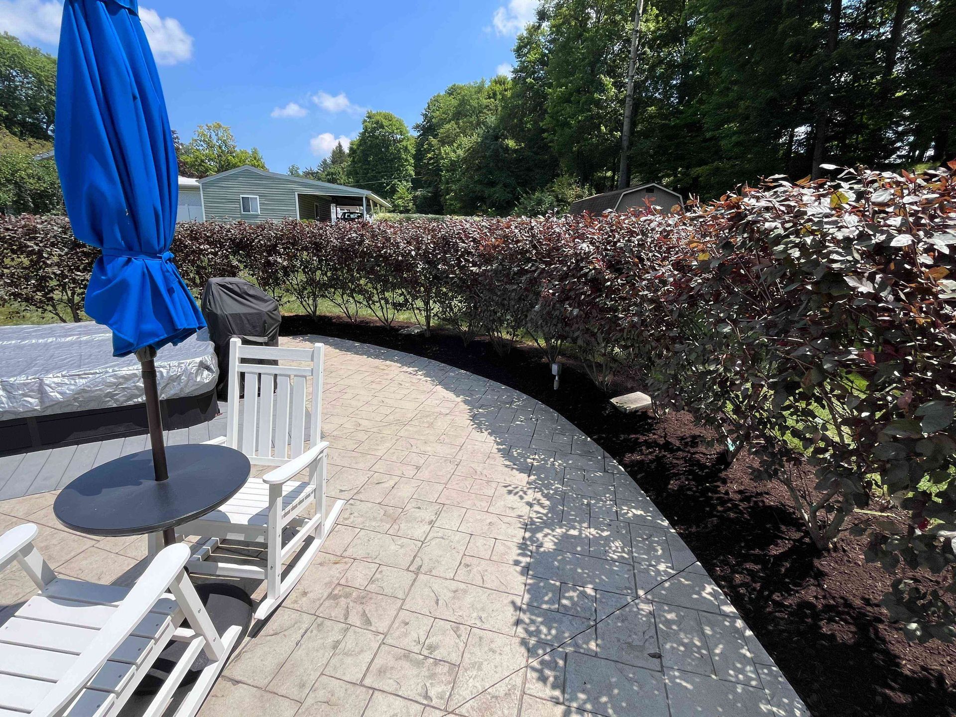 A patio with a blue umbrella, white rocking chairs, and a curved brick pathway lined by a dark burgundy hedge.