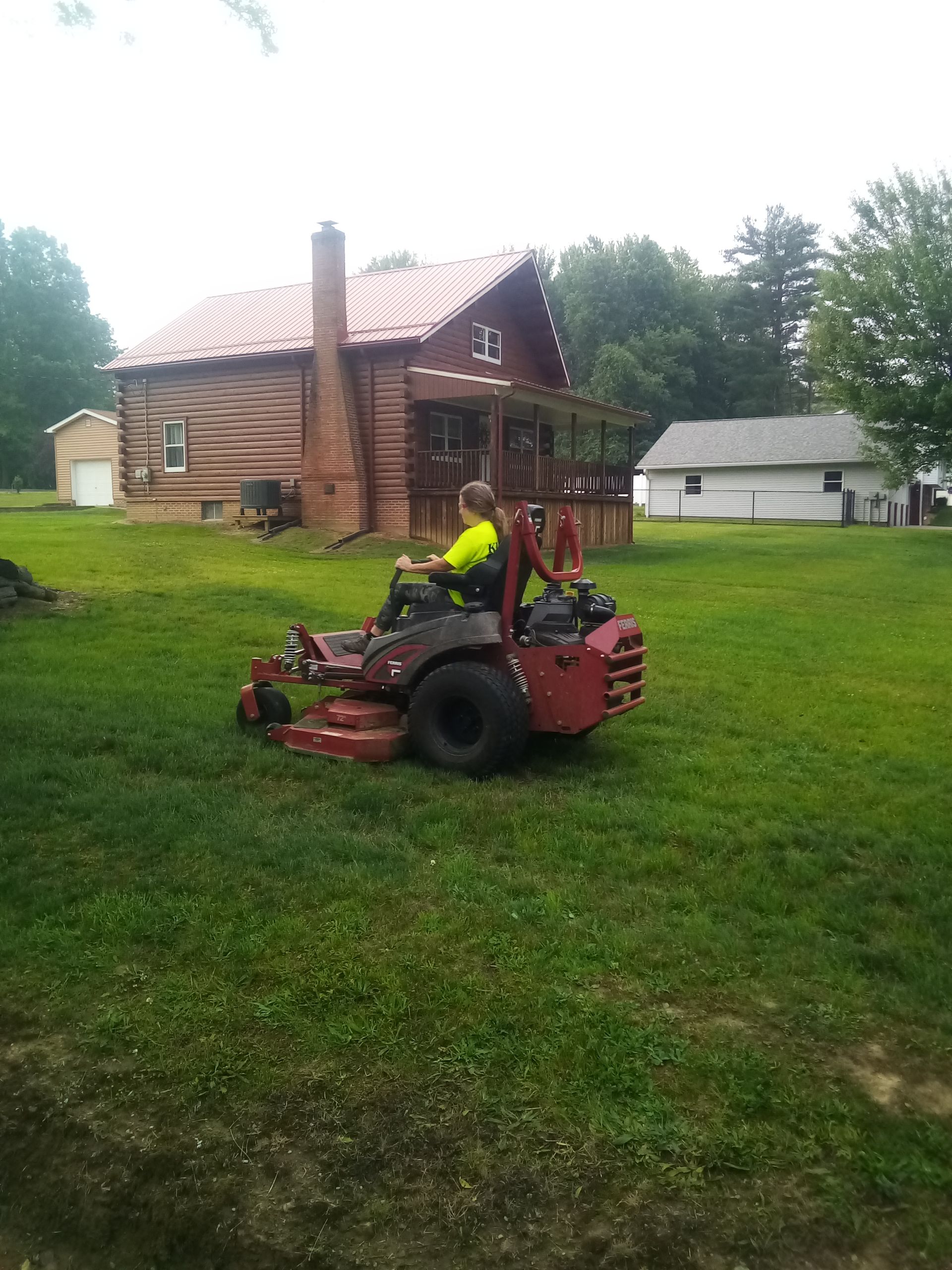 A woman is riding a lawn mower in front of a log cabin.