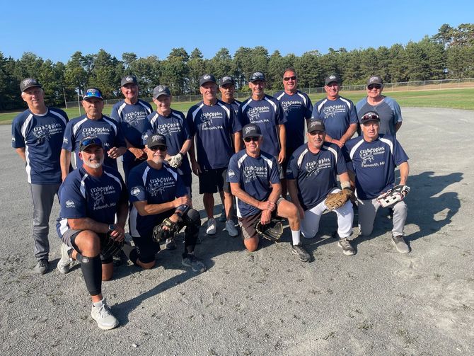 A team of fifteen softball players in matching navy jerseys and caps pose for a photo on a dirt field on a sunny day.