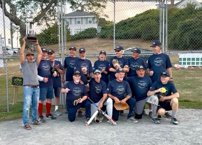 A team of softball players wearing matching dark shirts poses for a photo on a dirt field, holding a trophy and baseballs.