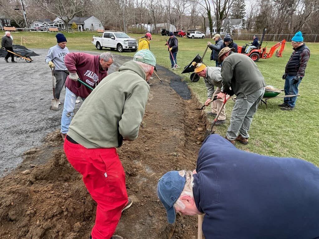 A person wearing a hat and orange shirt, holding a baseball bat on their shoulder on a baseball field.