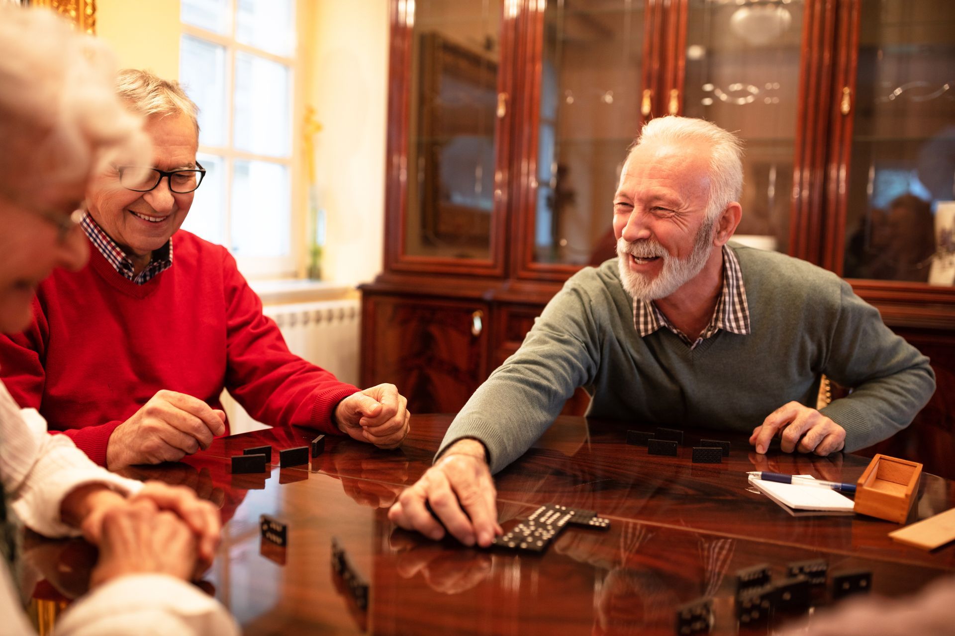 Three people sit around a polished wooden table in a dimly lit room, smiling while playing a game of dominoes.