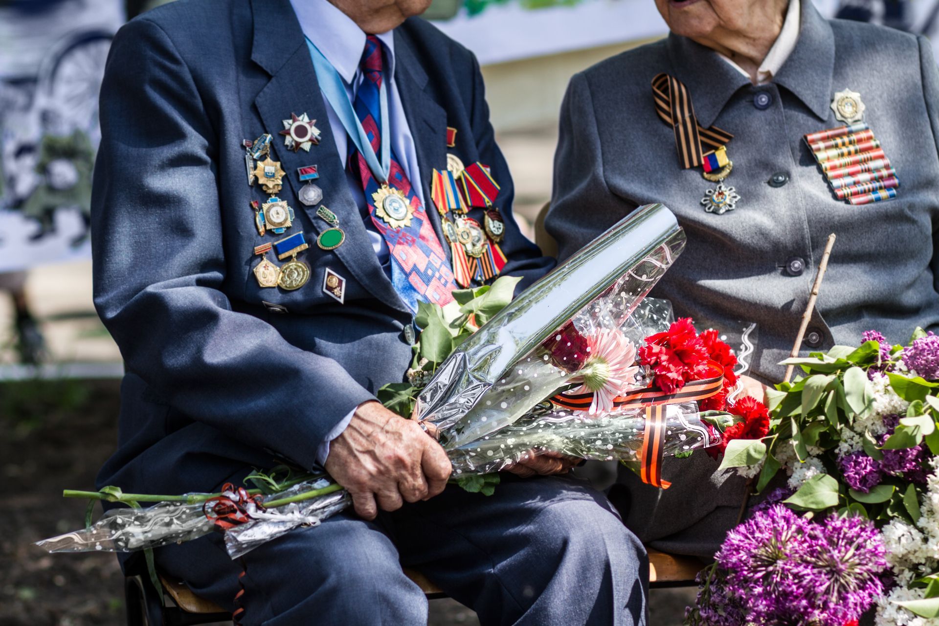 Two veterans in uniforms covered in military medals hold bouquets of flowers and red carnations.