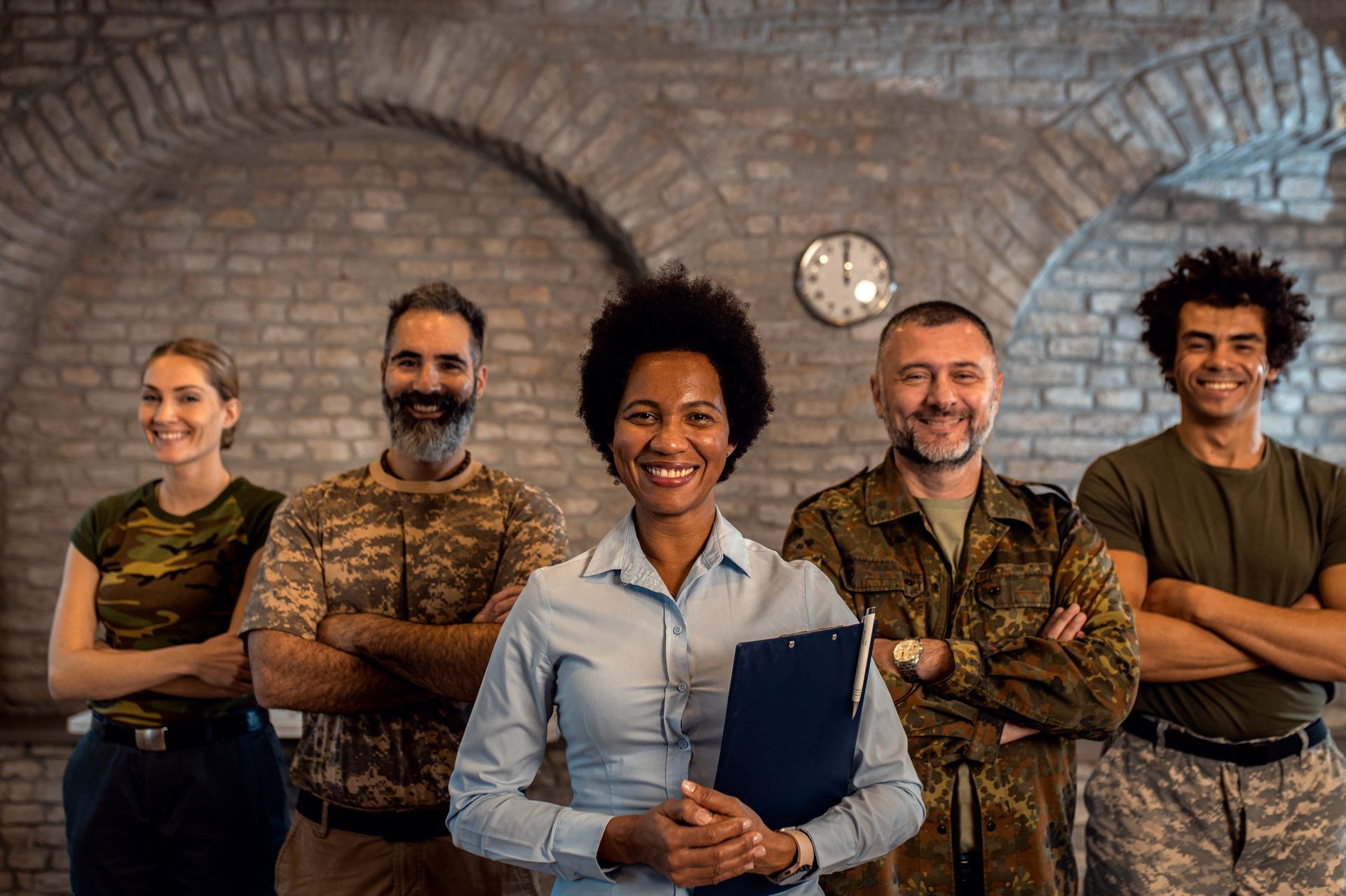A group of people, some in camouflage uniforms, standing together with arms crossed against a brick wall.