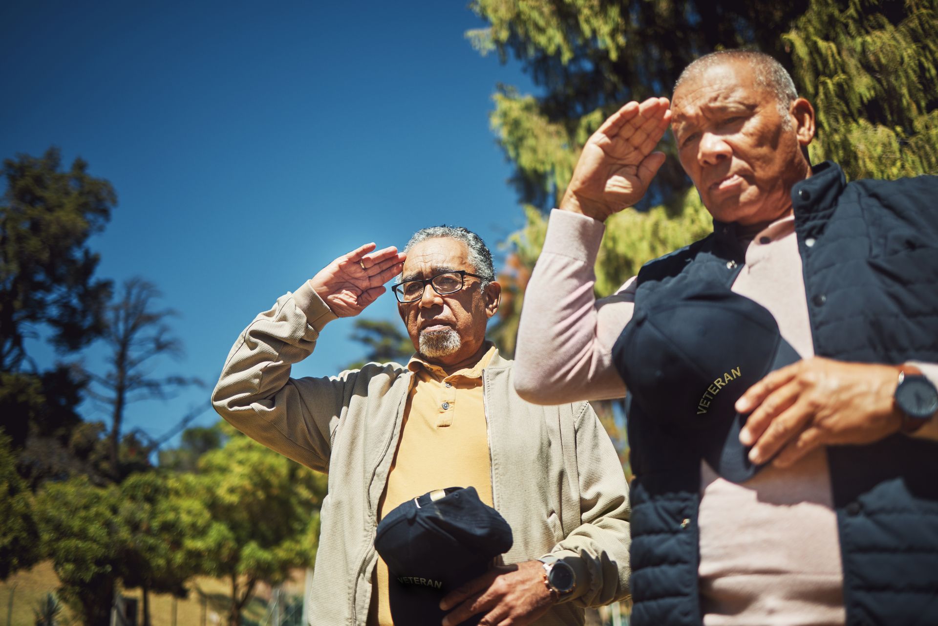 Two people standing outdoors, saluting with solemn expressions while holding hats.