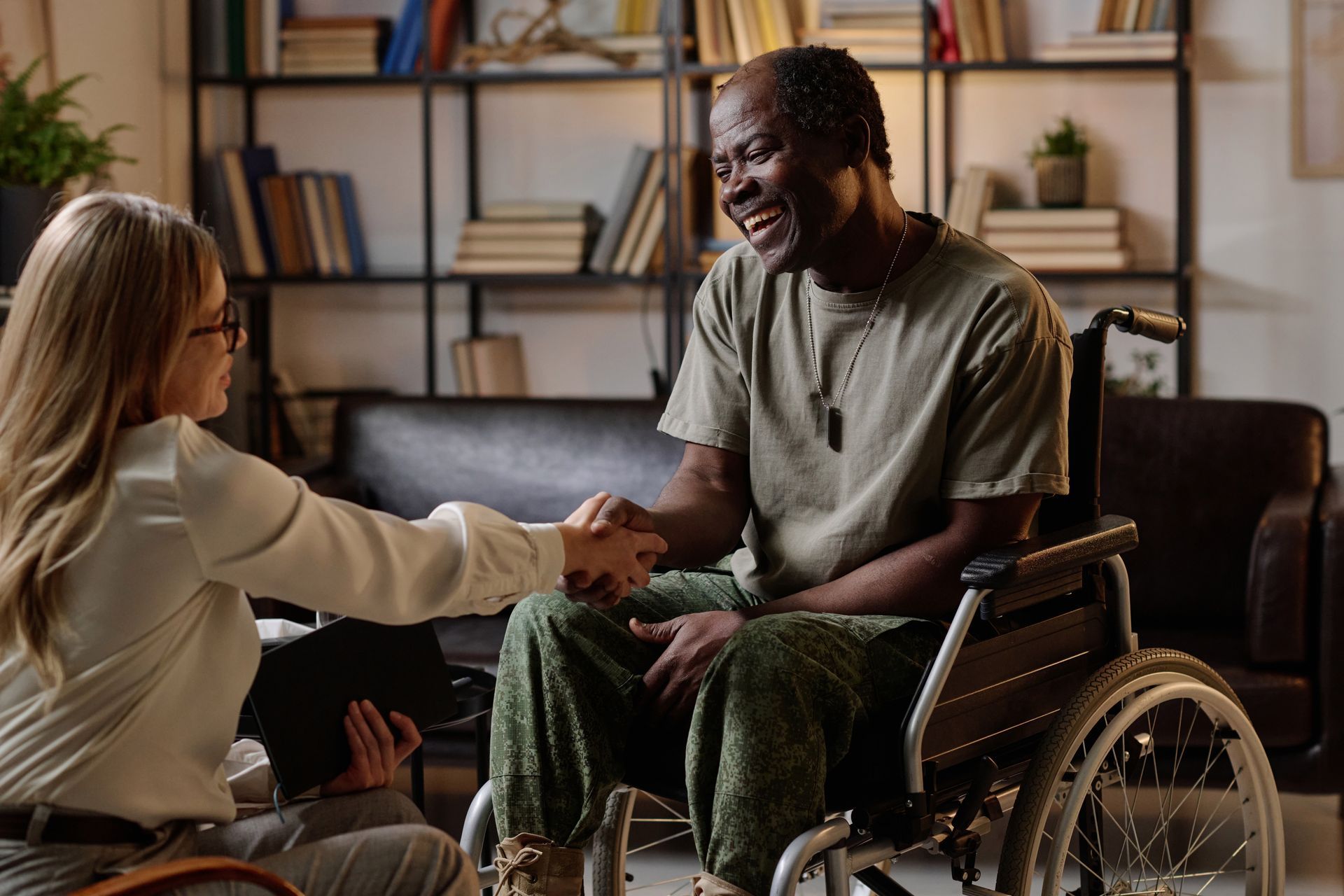 A person in a wheelchair shakes hands with a seated visitor in an office setting with shelves of books in the background.