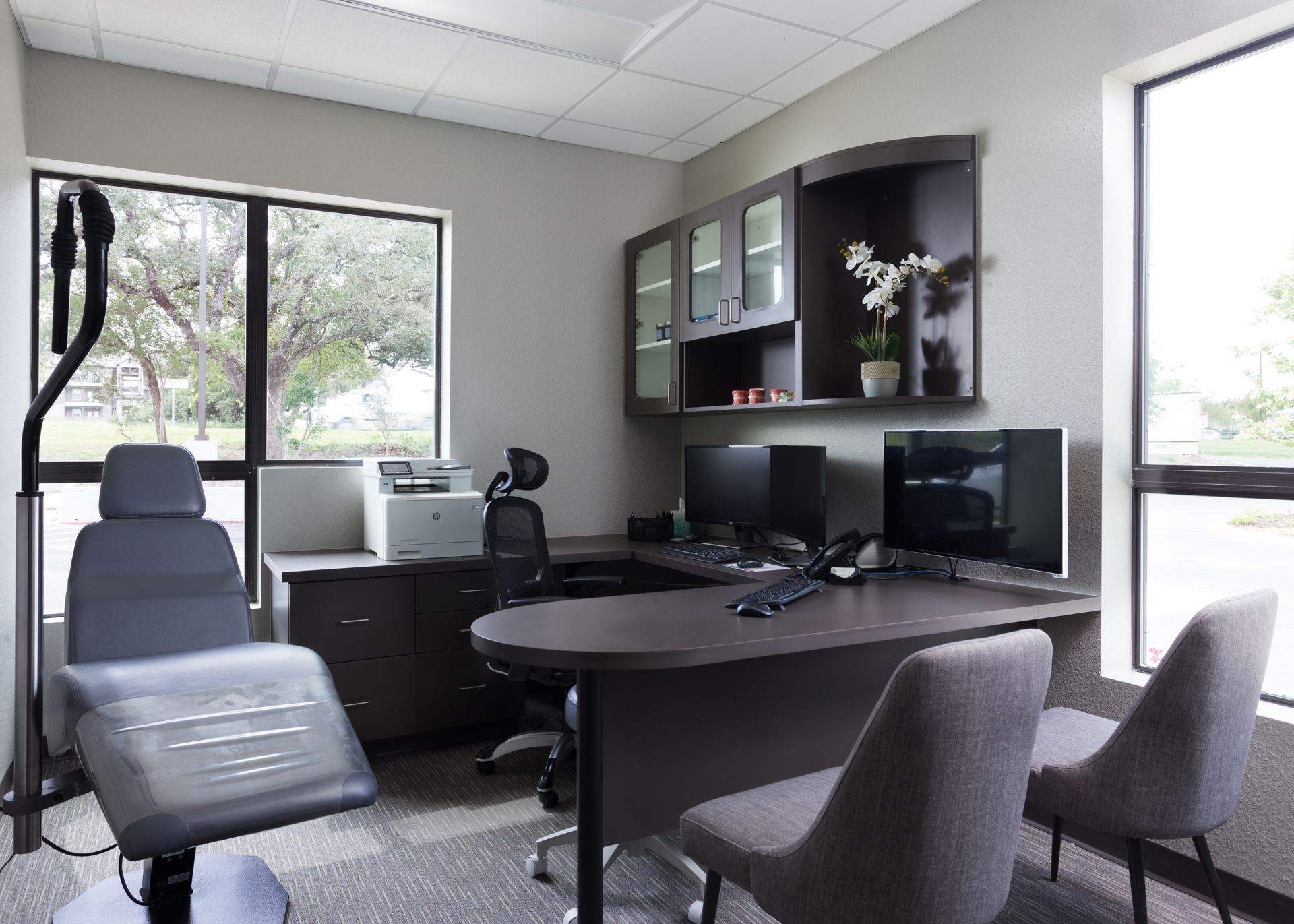 A professional office featuring a dark desk, two chairs, a computer, a printer, and a grey examination chair by a window.