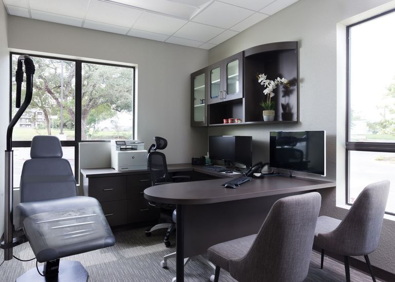 A professional office featuring a dark desk, two chairs, a computer, a printer, and a grey examination chair by a window.
