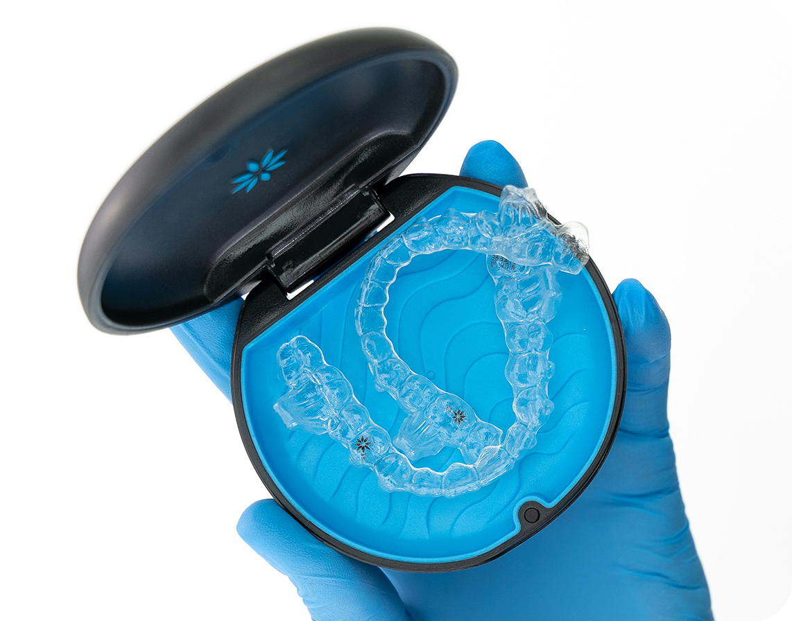 A hand in a blue medical glove holds a round black and blue case containing a clear orthodontic aligner.