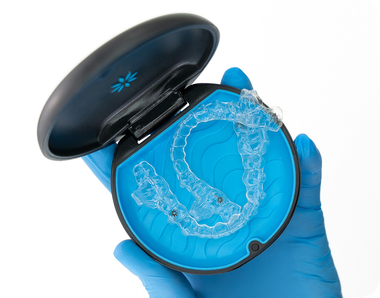 A hand in a blue medical glove holds a round black and blue case containing a clear orthodontic aligner.