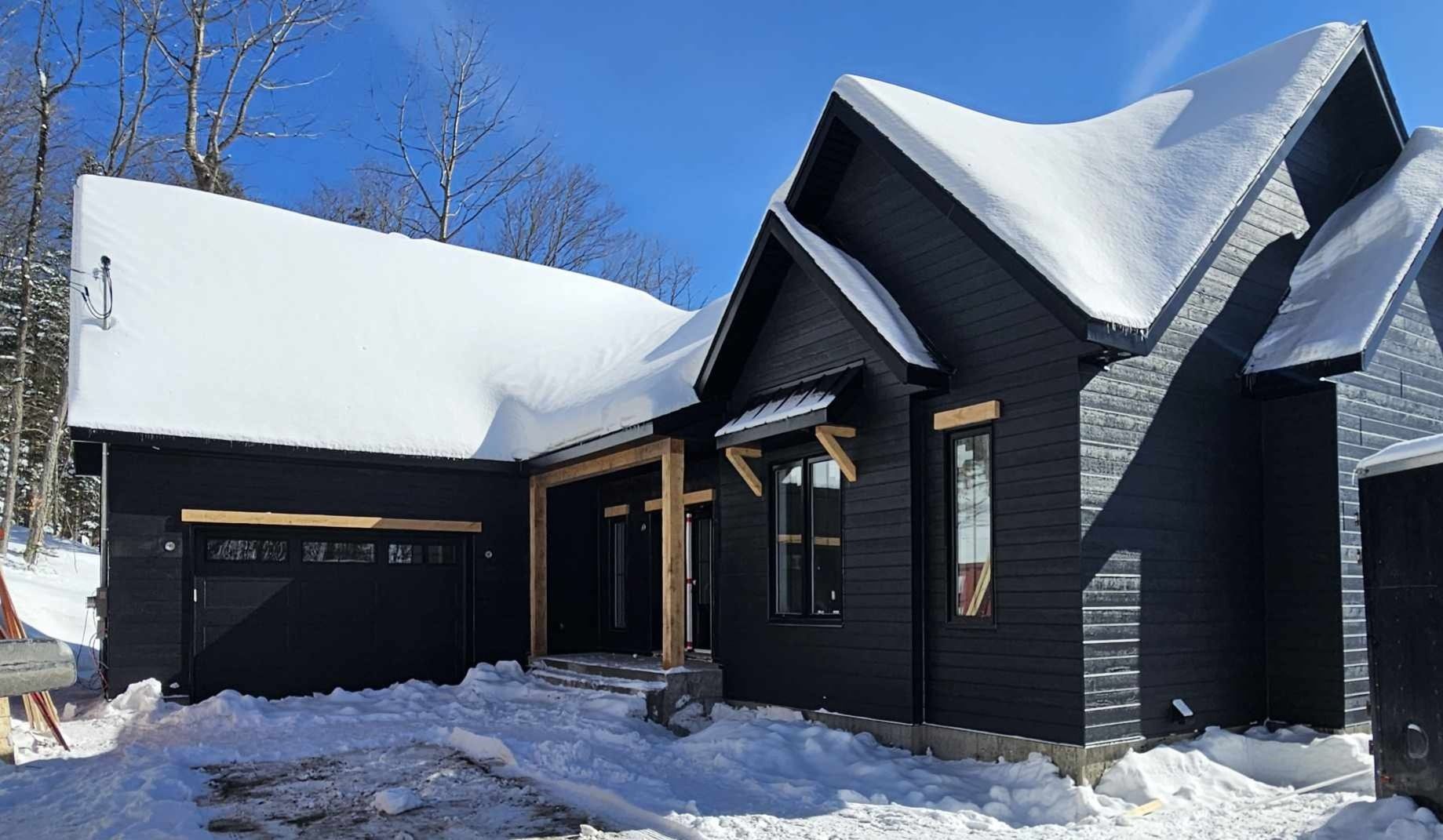 Une maison noire est recouverte de neige par une journée ensoleillée.