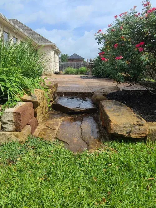 Stone water feature in a backyard, with a patio, flowers, and a house. Green grass and blue sky.