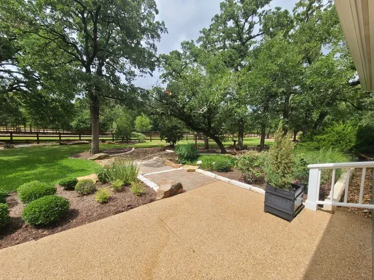 Exterior view: tan patio with greenery, trees, and a grassy lawn.