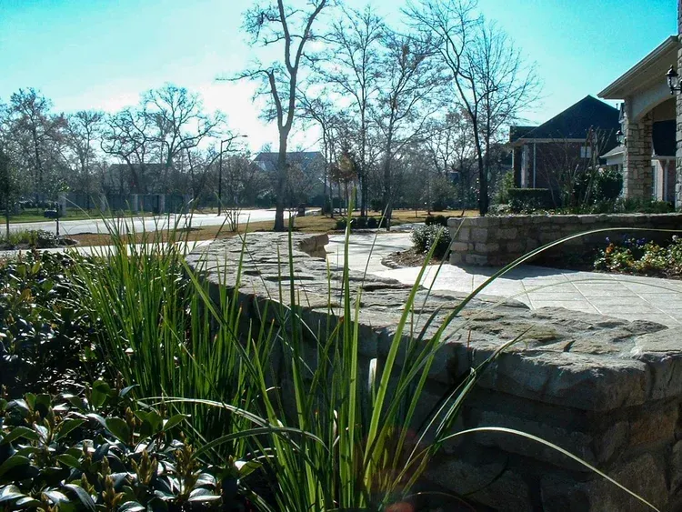 A stone retaining wall with tall green grass, next to a house and street with leafless trees under a blue sky.
