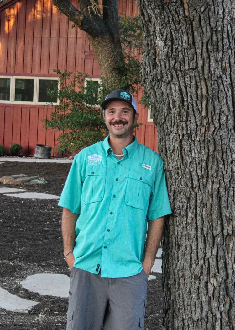 Man in light blue shirt and khaki pants, leaning against tree, red barn in background.