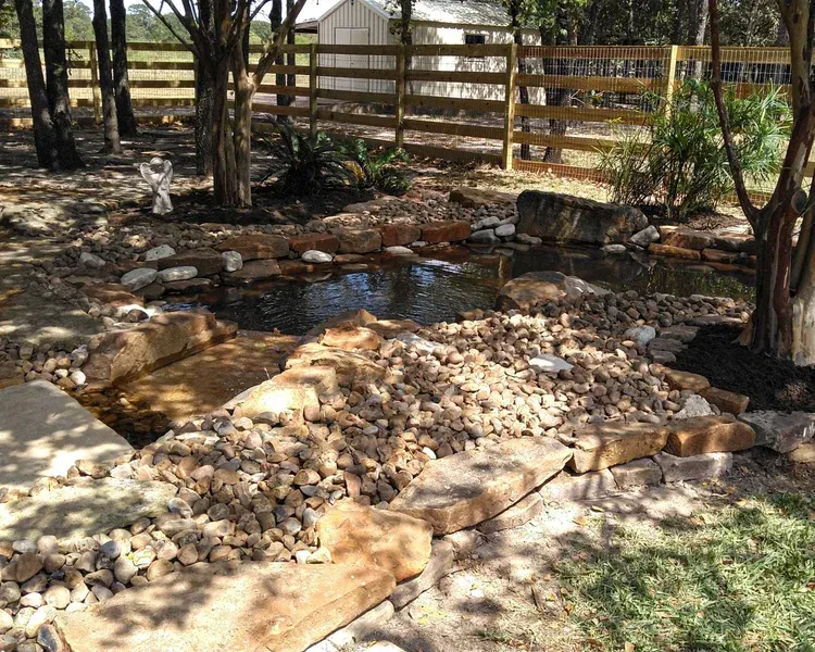 Pond surrounded by rocks and landscaping, near a wooden fence and trees.