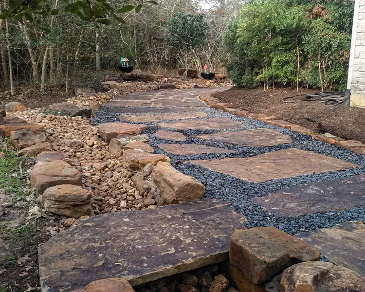 Stone pathway through a wooded yard, bordered by rocks and gravel, leading to trees.