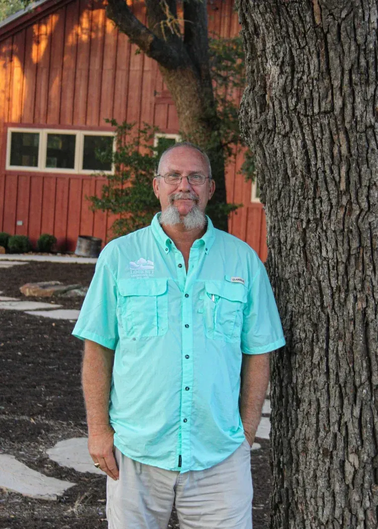 Man in light blue shirt and khaki pants, leaning against tree, red barn in background.