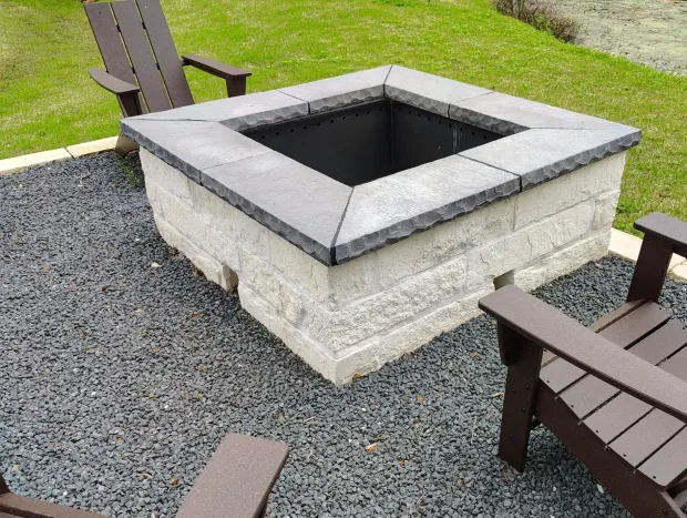 Square stone fire pit with dark gravel, two brown chairs, and green grass.