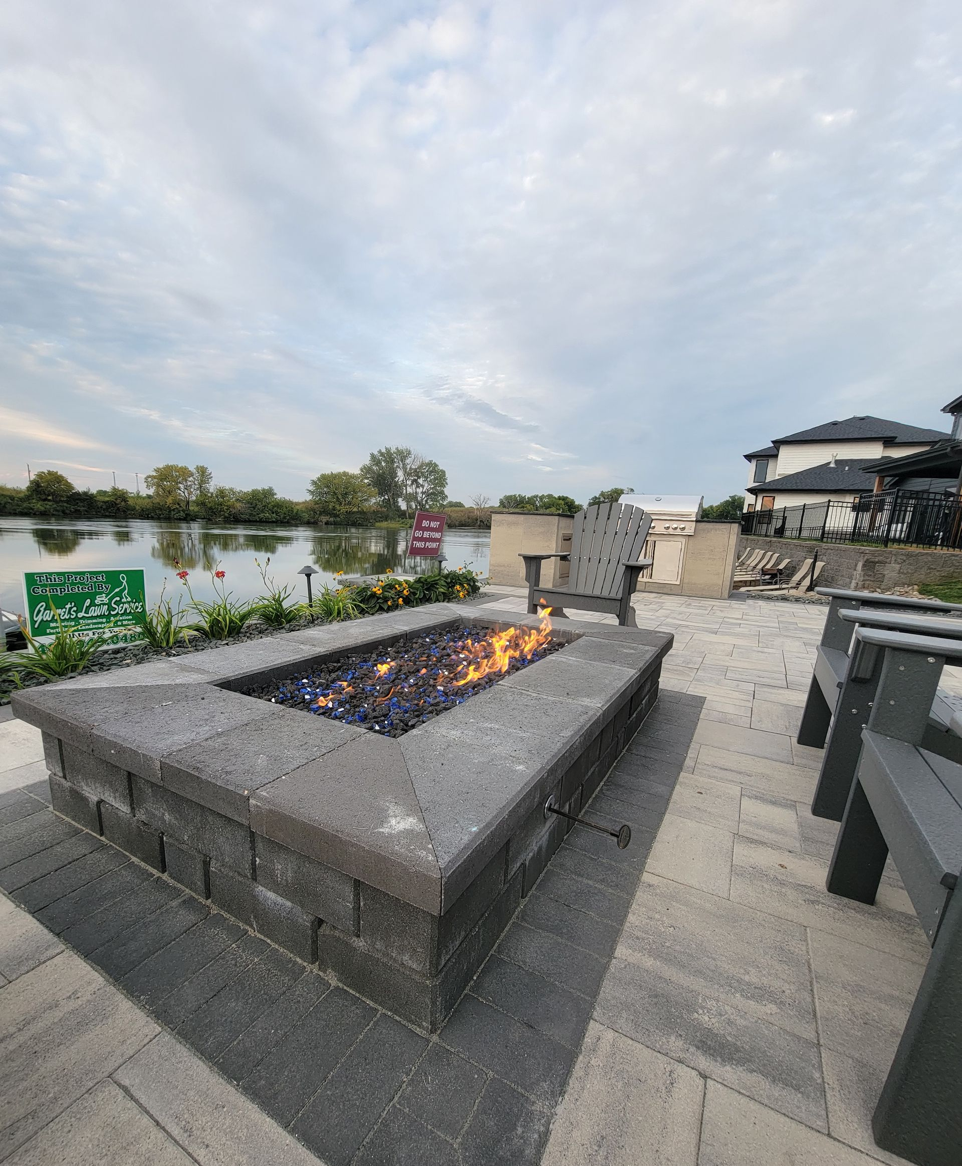 Fire pit with blue flames, surrounded by chairs, on a patio overlooking a lake.