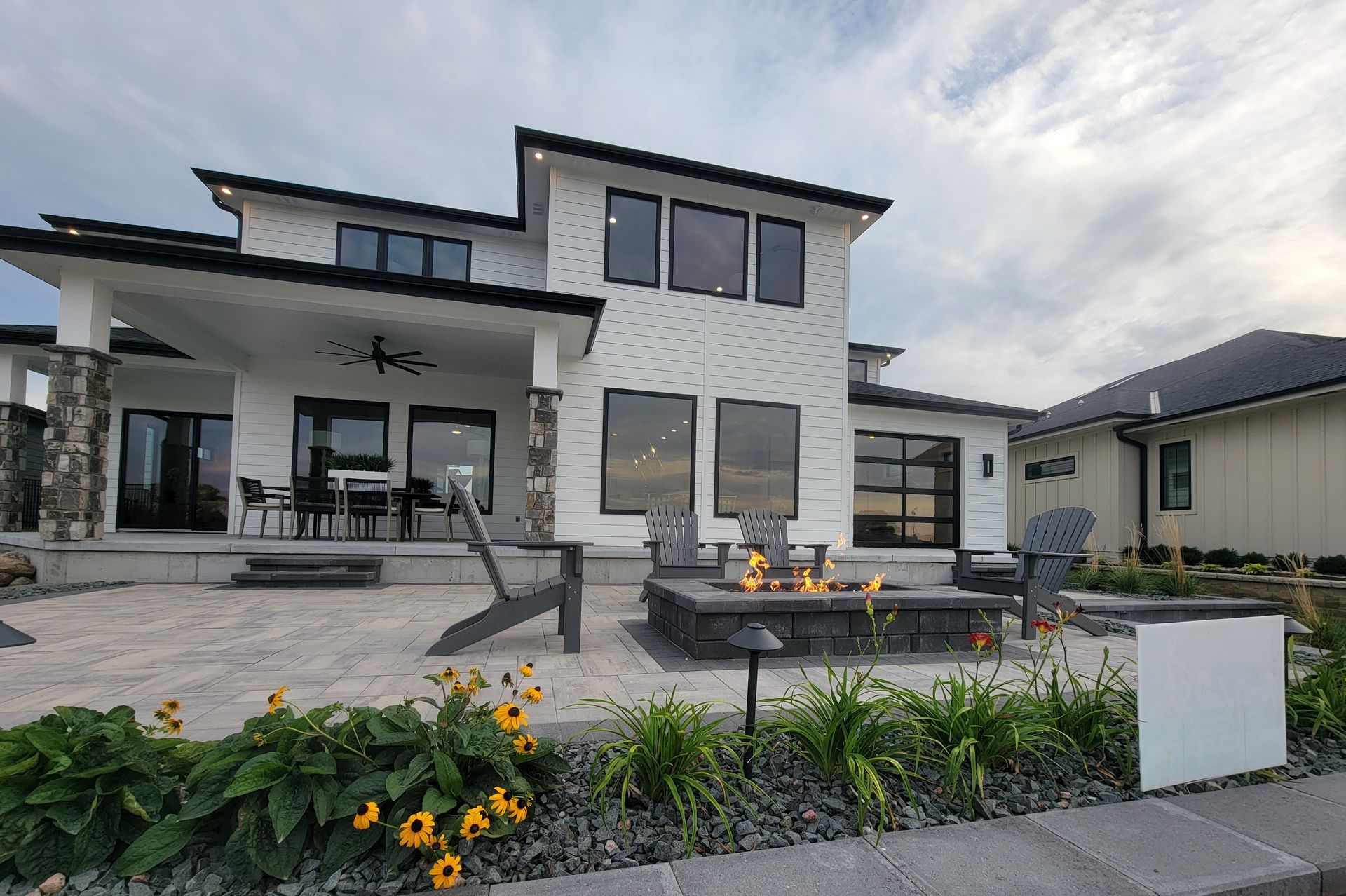 Modern white house with patio, fire pit, and flowers; cloudy sky.