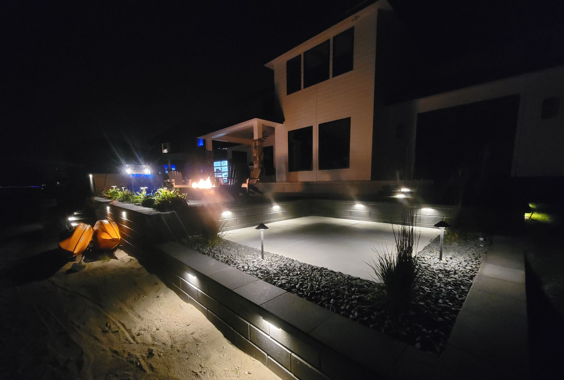 Nighttime view of a modern home with a lit patio, landscaping, and fire pit.