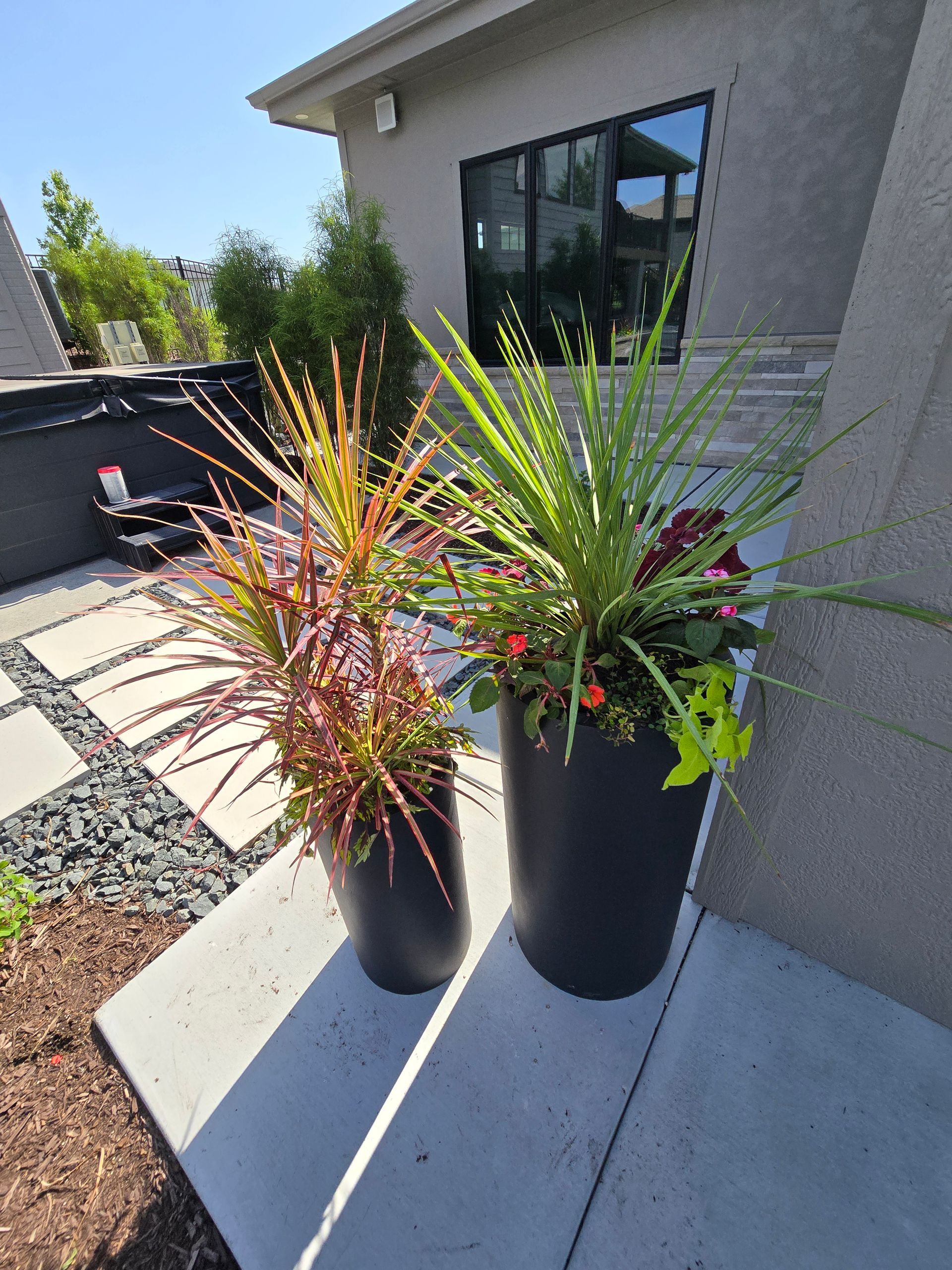 Two black planters with colorful plants on a patio next to a building.