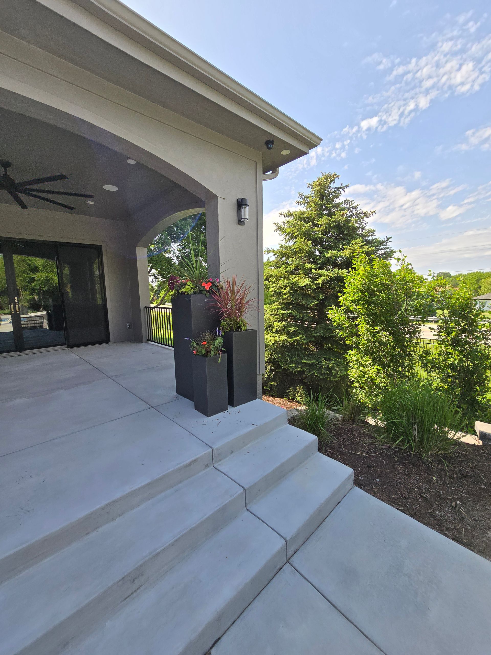 Grey concrete patio with steps, planters with flowers, and trees on a sunny day.