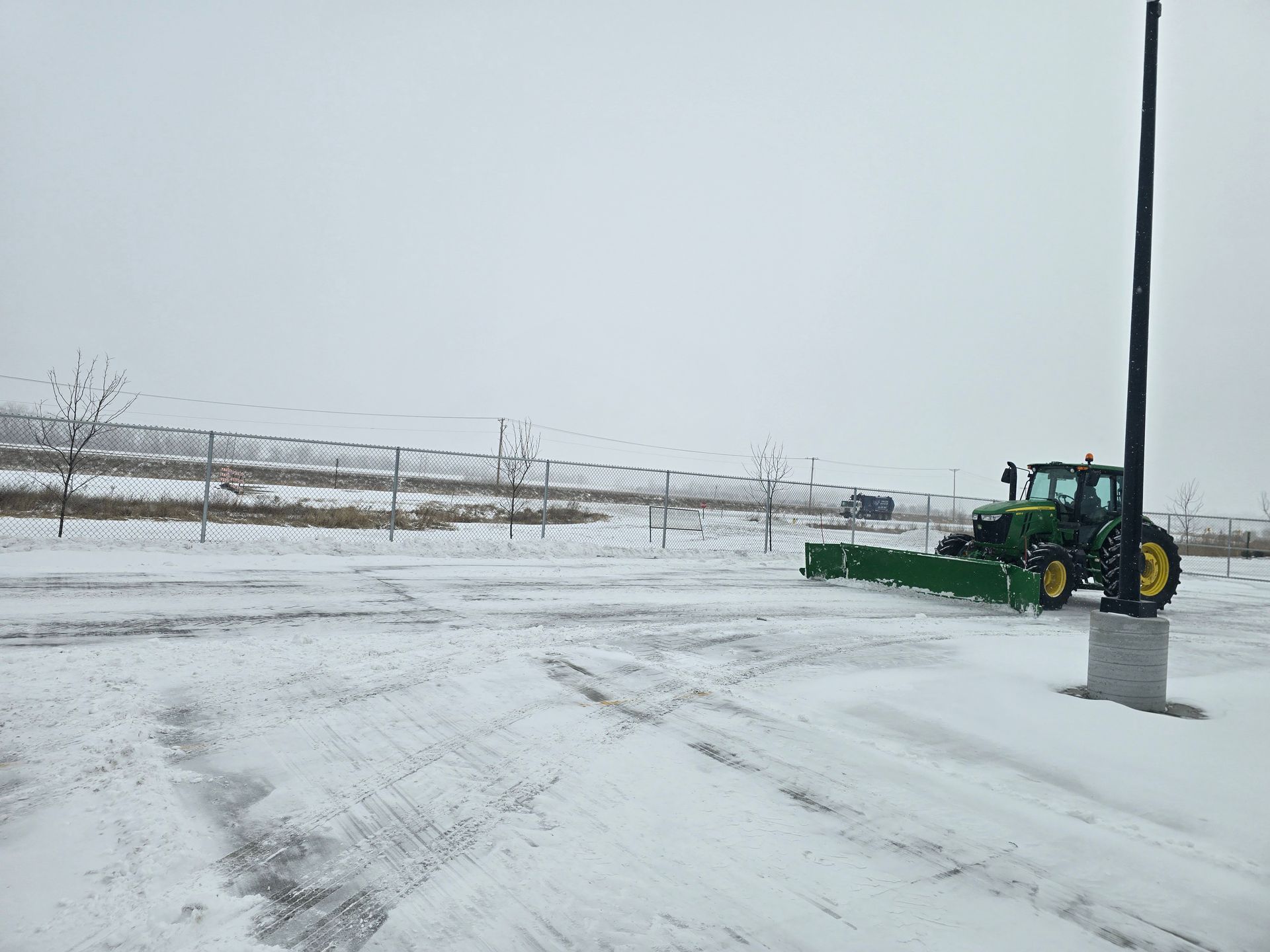 A green tractor plowing snow in a parking area on a cloudy day.
