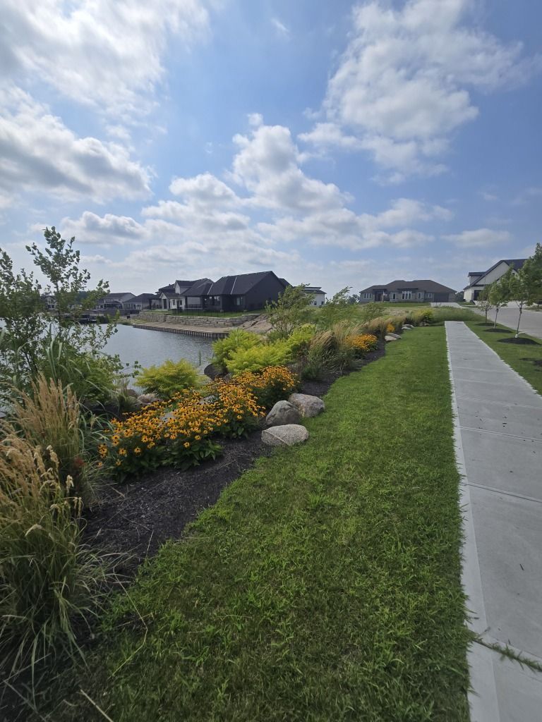 Lush landscaping along a lake with sidewalk and houses under a cloudy blue sky.