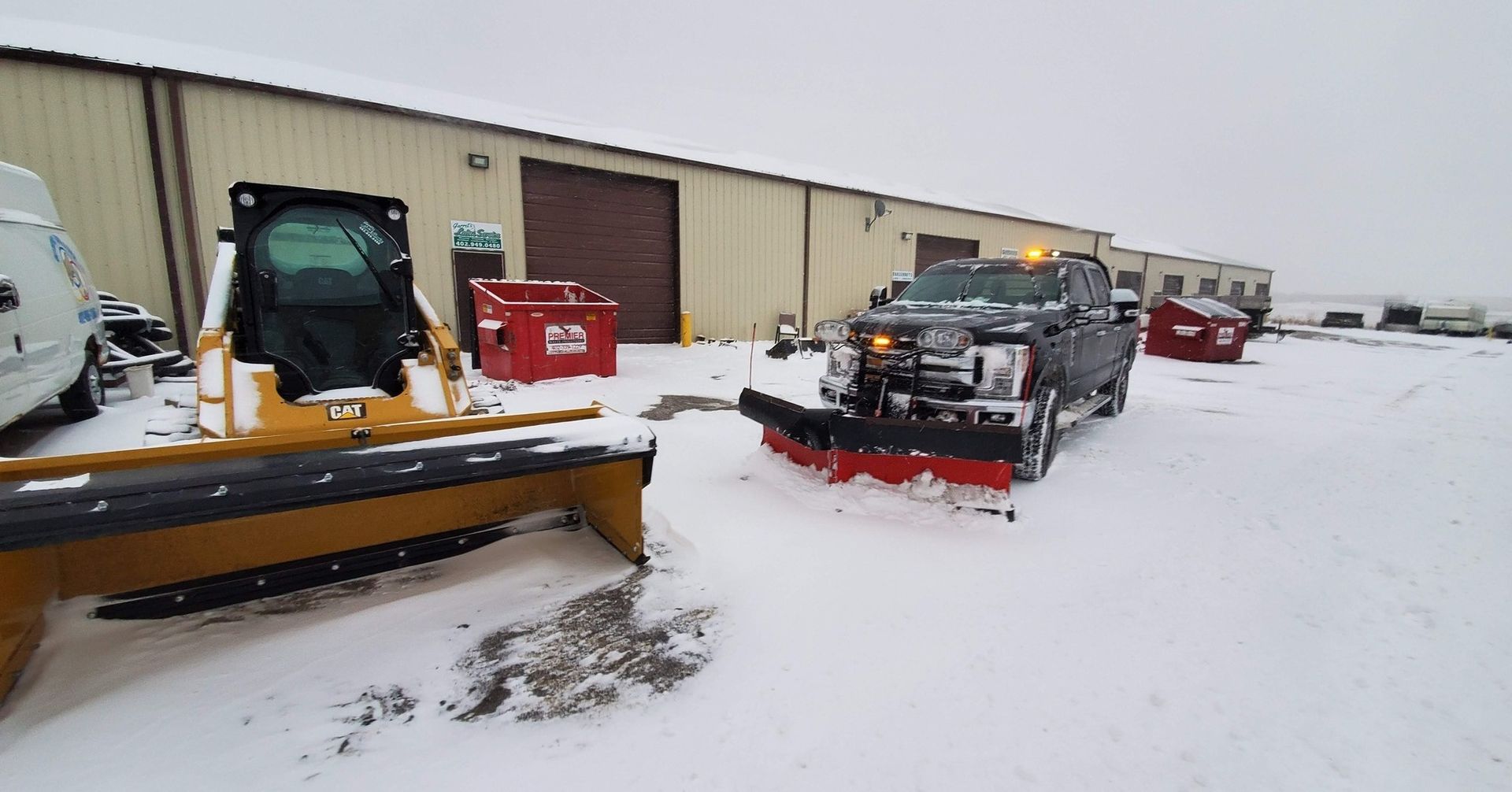 Snowplow truck and tractor with snowplow blades in front of a warehouse, snowy scene.