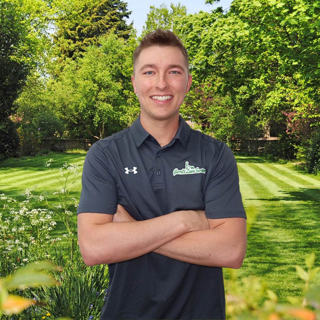 A man is standing in a garden with his arms crossed and smiling.