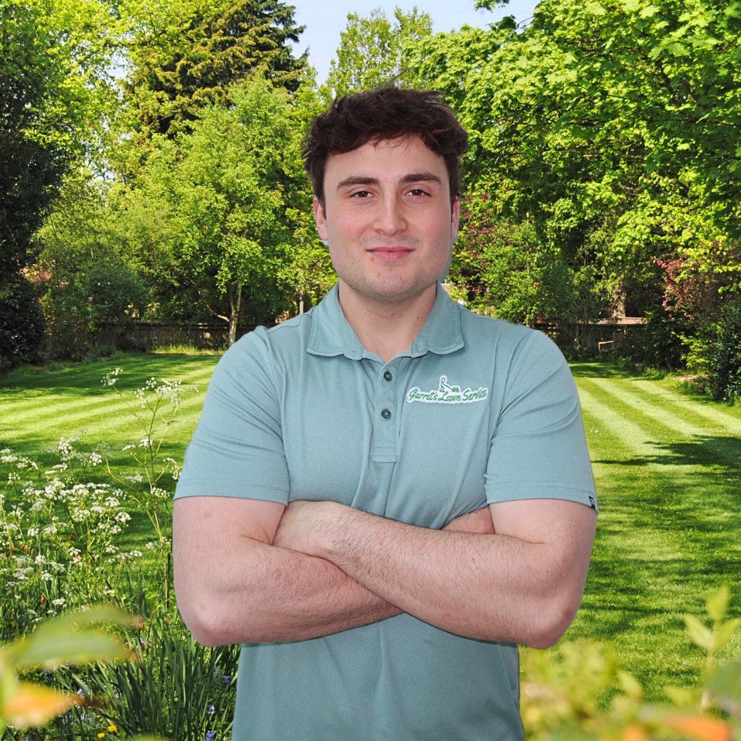A man in a blue shirt is standing with his arms crossed in a garden.