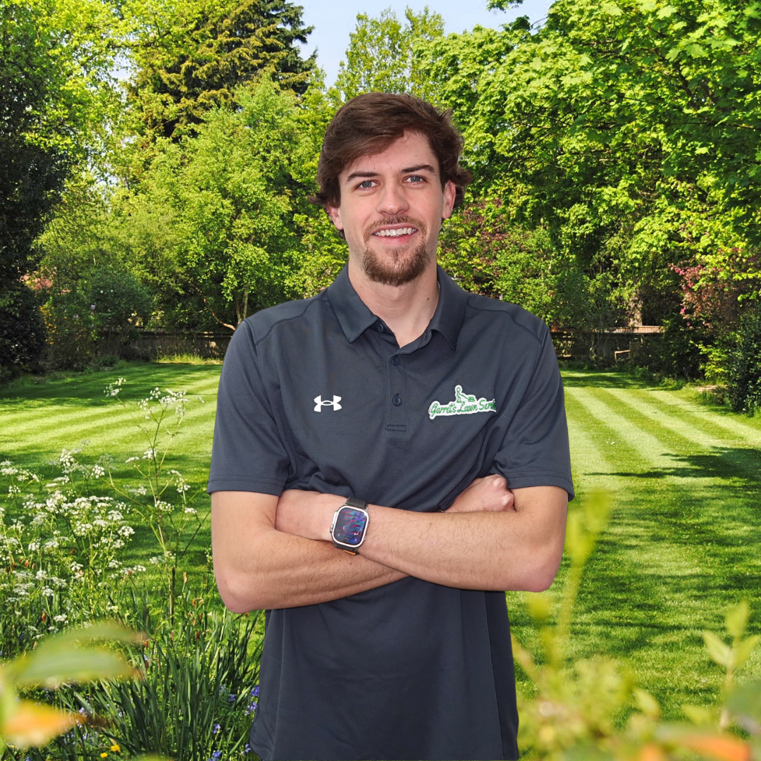 A man in a black under armour shirt is standing in a garden with his arms crossed.