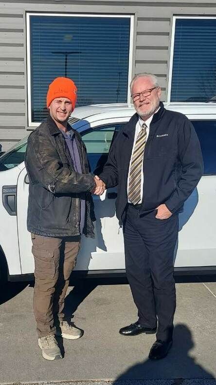 Two men are shaking hands in front of a white car.