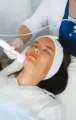 A technician uses a handheld cosmetic device to perform a facial treatment on a person lying down with a headband.